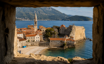 Pogled na Stari grad Budve kroz kameni otvor u zidinama, sa tirkiznim Jadranskim morem, srednjovekovnim zidinama i crvenim krovovima u sunčanom danu.