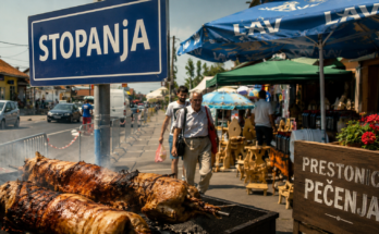 Tabla sa natpisom „Stopanja” na ulazu u selo poznato kao prestonica pečenja u Srbiji, sa pečenjarama i glavnom ulicom u pozadini.